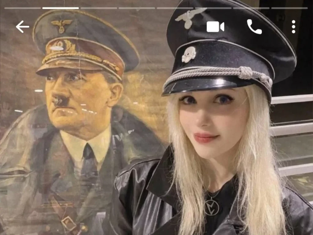 Marina Seren wearing a Nazi outfit, posing in front of a painting of Adolf Hitler.